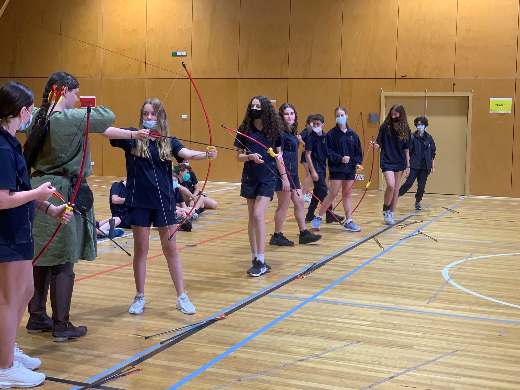 Senior School students are in a line in the gym, they are holding red bows and are about to shoot arrows - they are learning archery