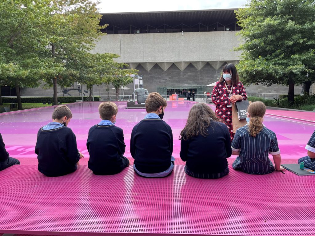 Five students have their back to the camera. They are at the pink lake at the NGV on an art excursion