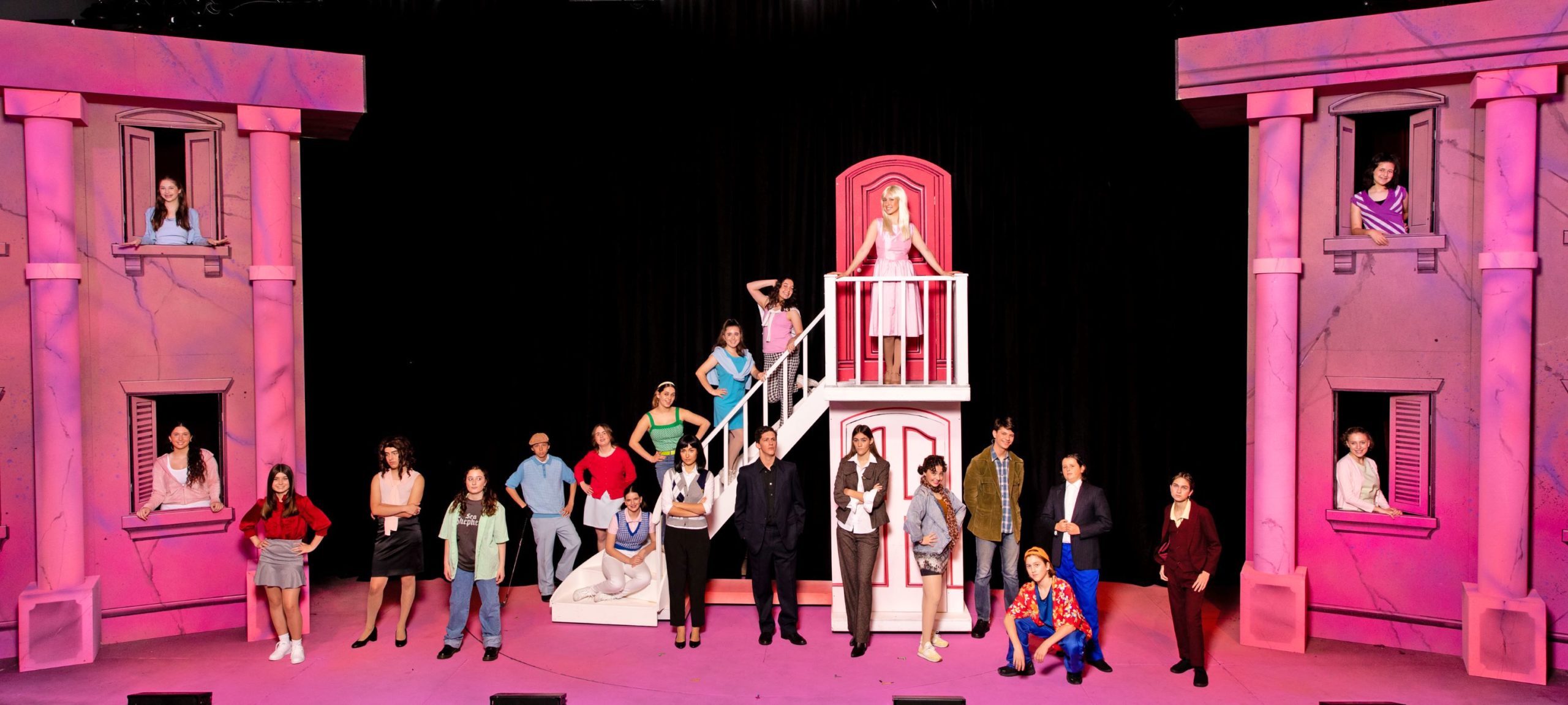The cast of Legally Blonde stand facing the camera, in costume, descending a staircase with Elle Woods at the top