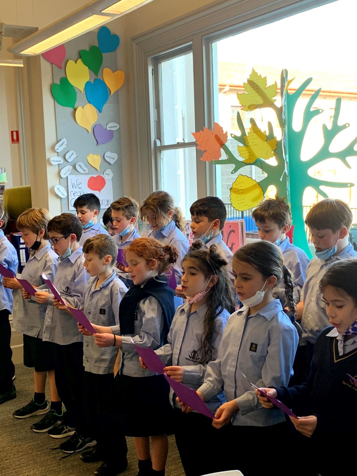 A group of Year 3 students are in the library. They are standing, reading a poem together aloud.