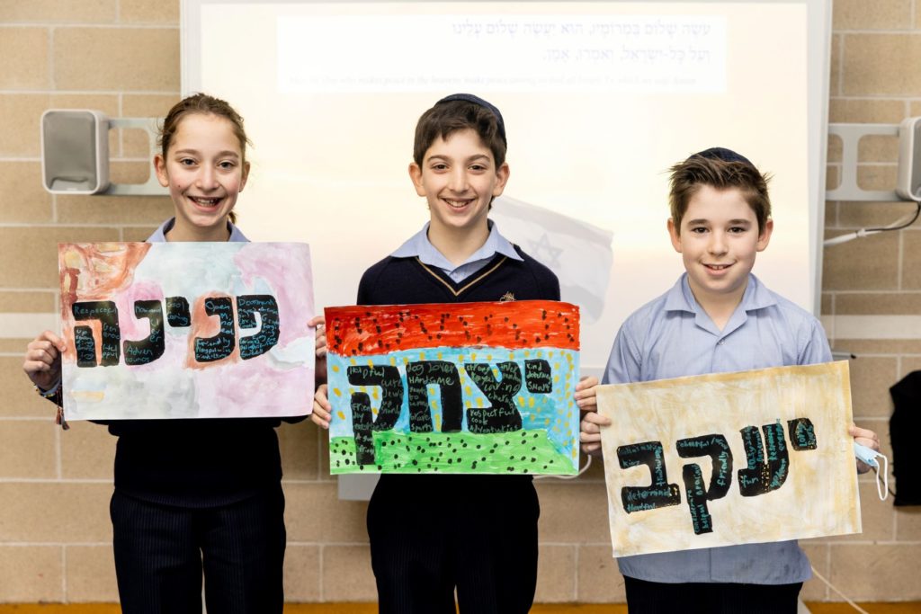 Three students are smiling looking at the camera. They hold paintings of their Hebrew names