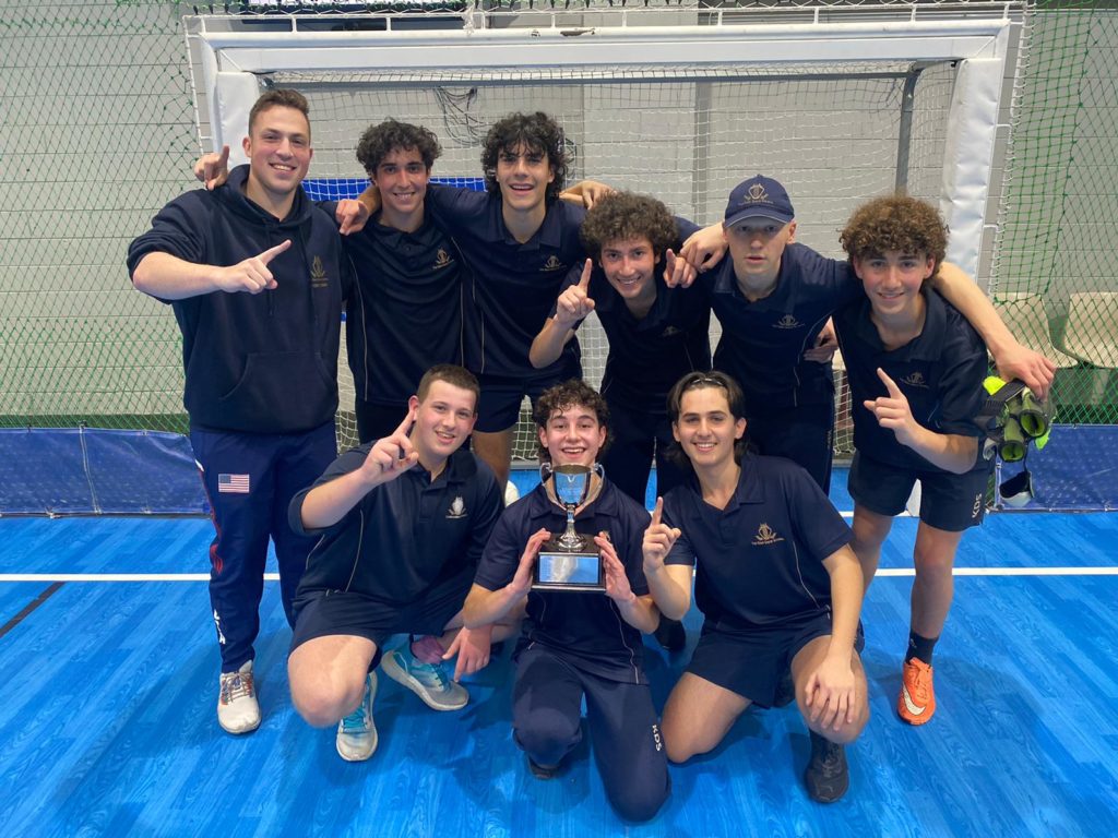 The boys soccer team is huddled together, they are looking at the camera and smiling. The boy crouching down in the middle is holding a trophy.
