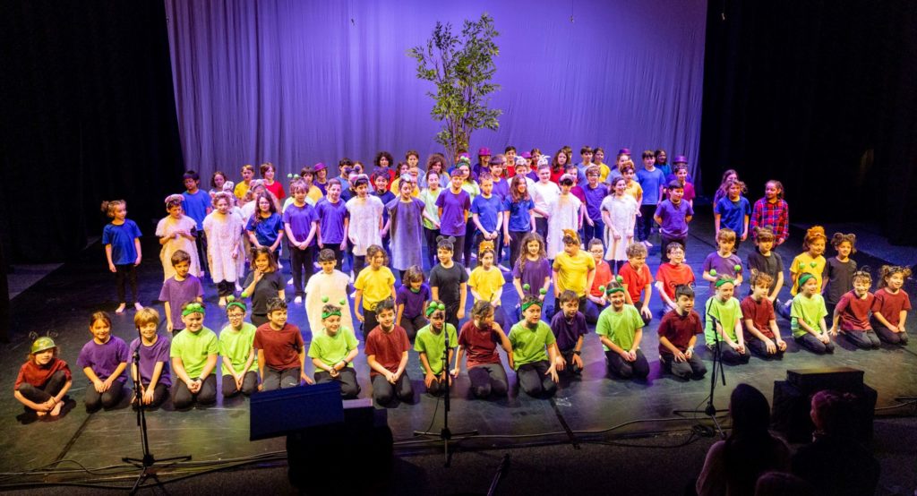 The Years 3 to 5 students are all in costume on stage with purple light and a real tree as the background. They have just performed their production