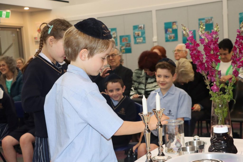 Two children are lighting Shabbat candles, there are children looking at the candles and grandparents sitting down.