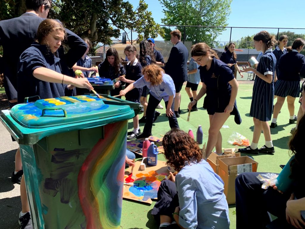 Photo shows a big rubbish bin being painted with rainbow swirls by students in uniform who are promoting sustainability
