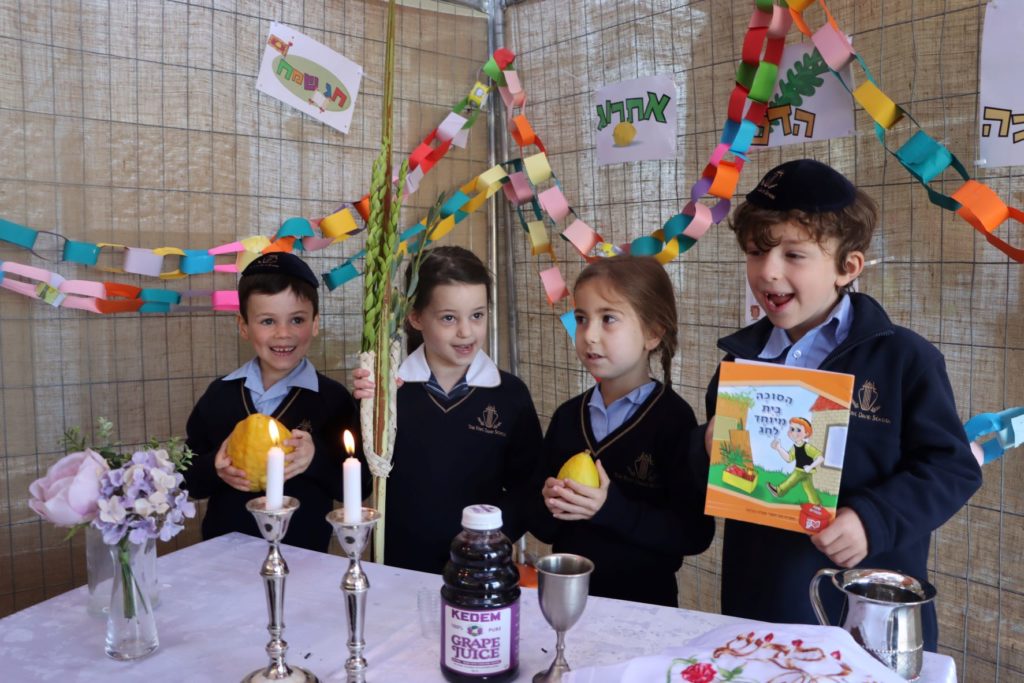 Four children are in the sukkah. Two are holding Etrogs. One holds a book in Hebrew about Sukkot,. There are Shabbat candles on the tables, along with grape juice for kiddush.