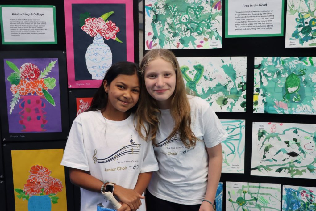 Two Year 5 students are looking at the camera and smiling. They are wearing KDS choir t-shirts. They are standing in front of colourful art painted by King David students.