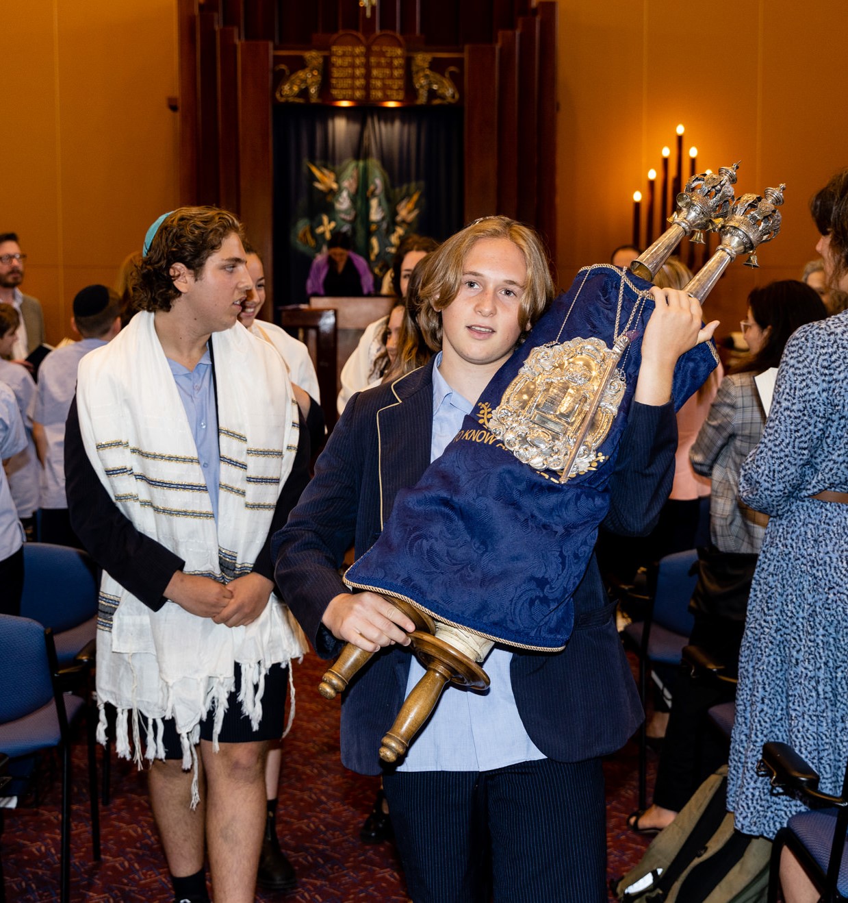 A male student holds a Torah decorated with a blue King David School Torah cover. He is looking at the camera. A male student beside him is wearing a white Talit. He is looking to the side.