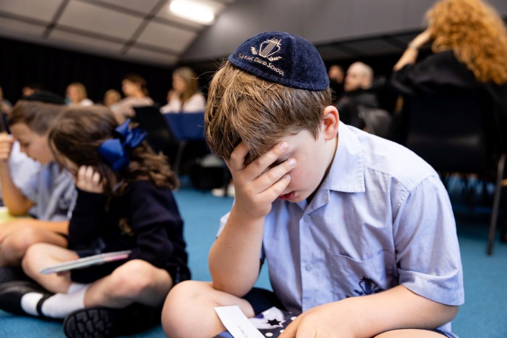 A student wears a King David School Kippah. He is covering his eyes with his hand and holding a new Siddur made for him