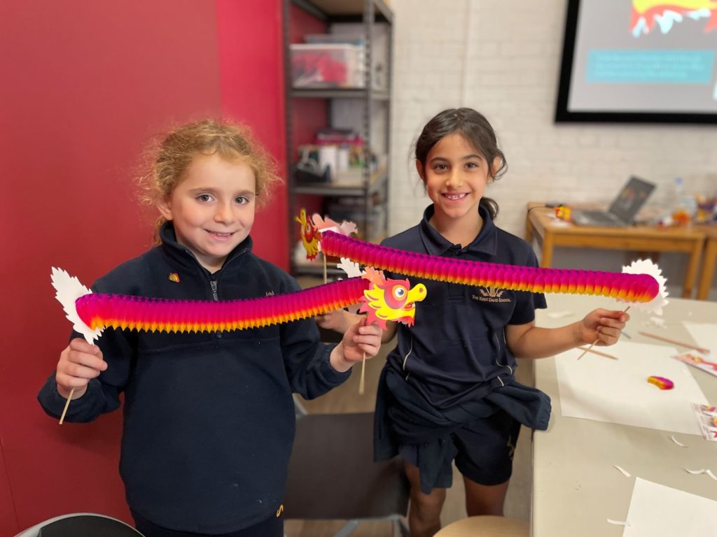 Two Year 2 students hold purple and red paper Chinese dragons they have made. They are at the Chinese Museum. They are looking at the camera and smiling.
