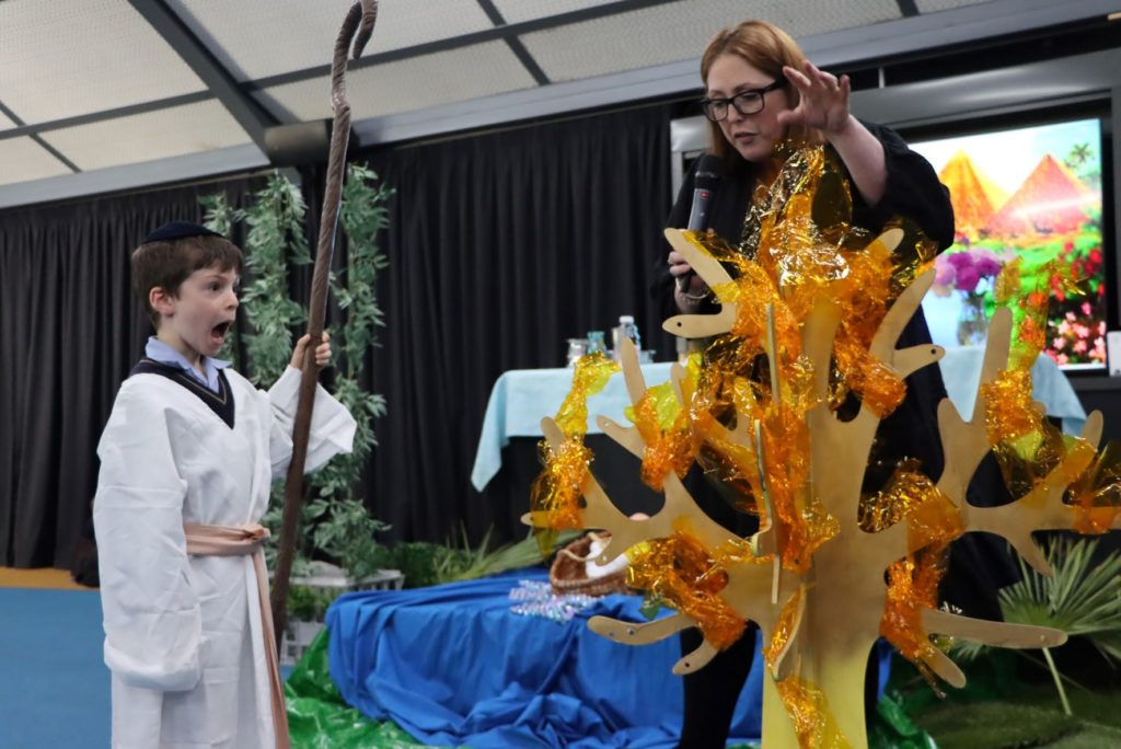 A Junior School student is dressed as Moshe when he was a shepherd. He is standing in front of a burning bush made of orange and yellow cellophane. He is about to hear God's voice.