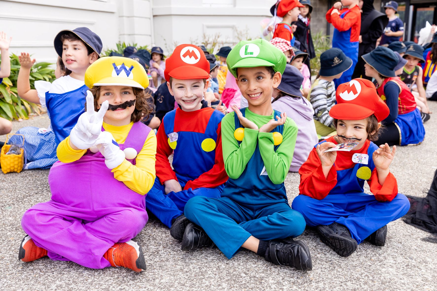 Four students are wearing costumes in the style of the Super Mario Brothers. They have fake mustaches and hats. They are sitting cross legged and are happy to be celebrating Purim at The King David School.