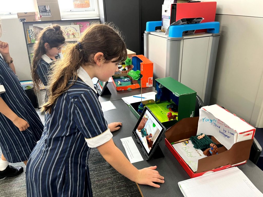 A Year 4 student stands in front of her laptop and stop motion figures. She is making a stop motion film.
