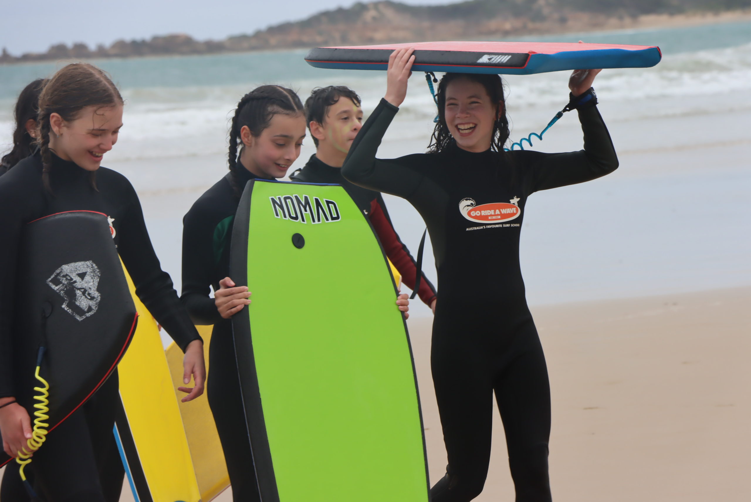 Five Year 7 students are on the beach. Two are holding green and yellow board boards against their bodies and one is holding a body board over their head. They are smiling and on the beach.
