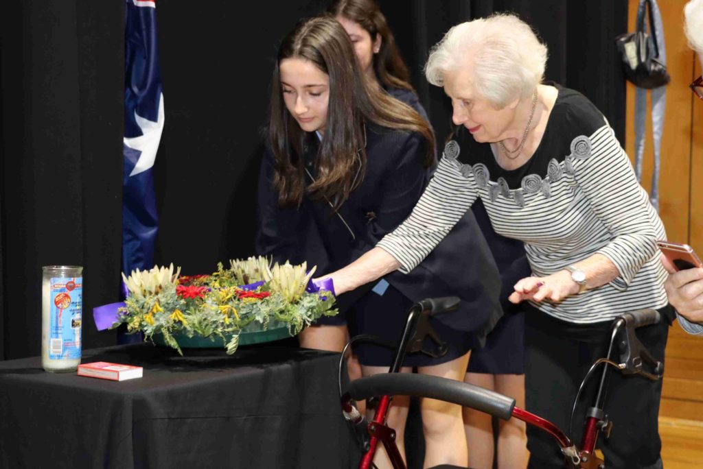 A student and her great grandmother together lay an ANZAC Day wreath on a table with a memorial candle. They stand in front of the Australian flag.