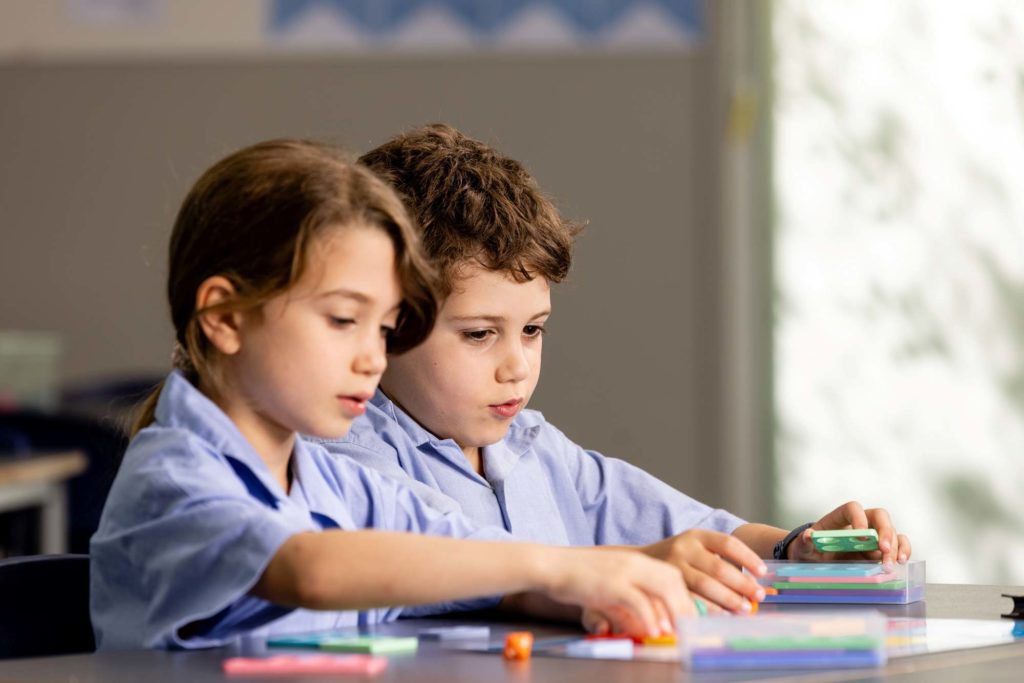 Two Junior School students are sitting working on a maths puzzle together. They are both holding pieces and looking down at the puzzle.
