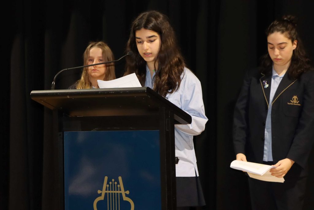 Three students stand solemnly at the Yom HaShoah assembly. One is speaking at the lectern.