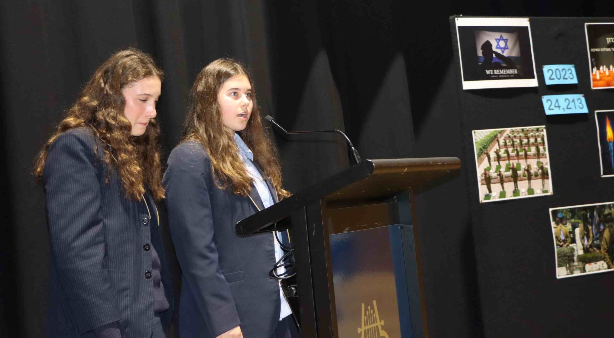 Two students are marking Yom HaZikaron. They look sad. One student looks down at the podium as she speaks.