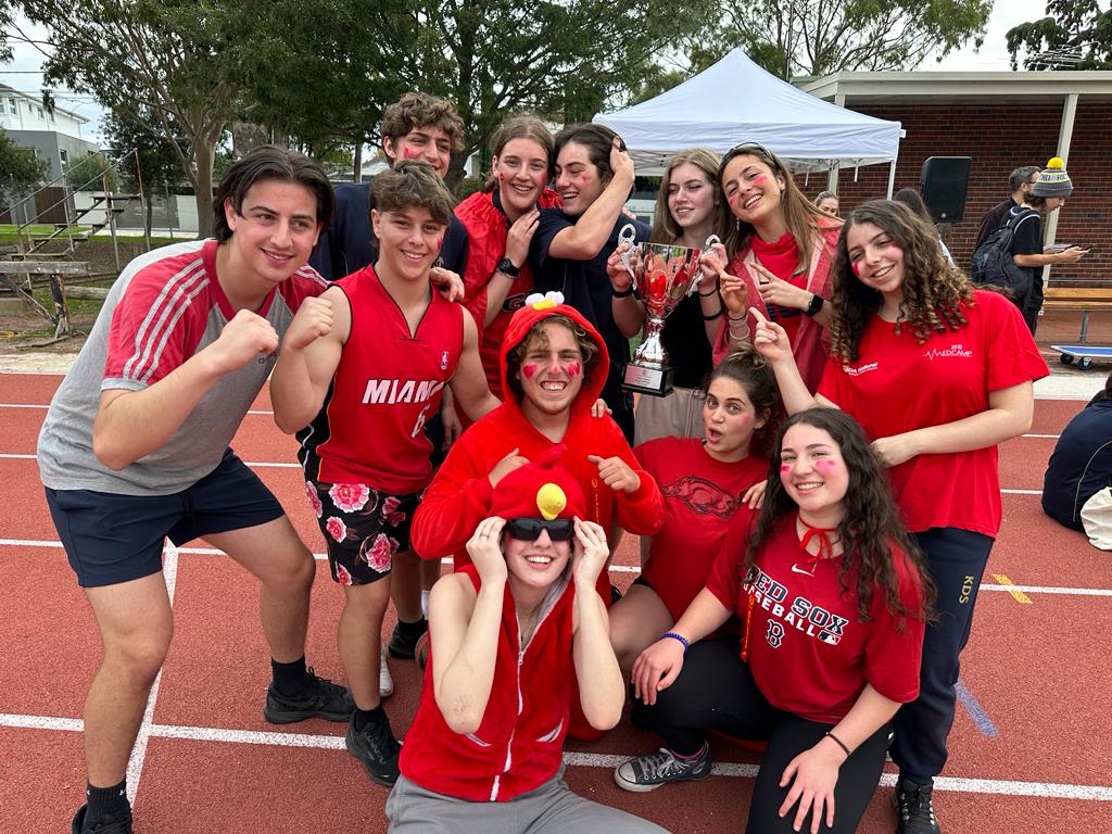 Year 12 members of Negev house smile and grin triumphantly after winning the 2023 Athletics Carnival. They wear red, the colour of Negev.