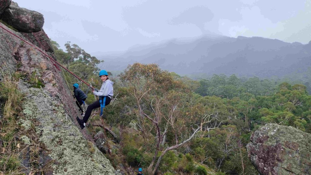A Year 8 student is abseiling up a cliff. She is wearing a bright blue helmet and looking at the camera, smiling.