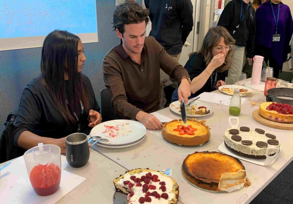 Three teachers are judging student made cheesecake for Shavuot. One is putting cheesecake in her mouth.