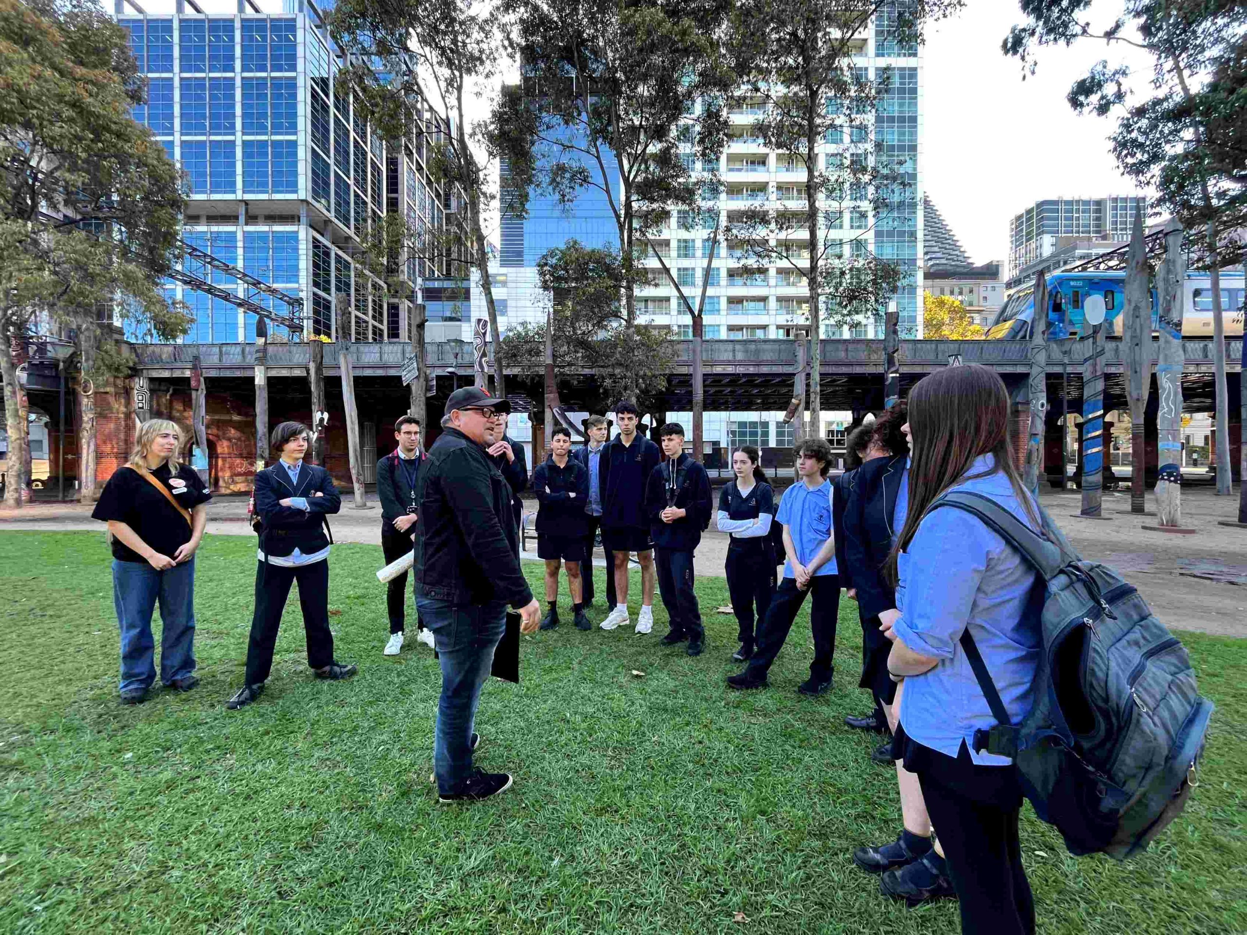 Year 10 students are on a First Nations walking tour with Dean Stewart, Wartha Worrung man in Melbourne's CBD. They have stopped on grass to listen to Dean.