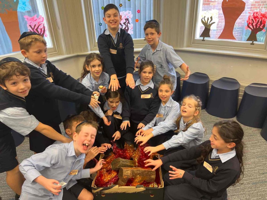 Junior School students warm their hands on a fire made out of cellophane as they celebrate Lag BaOmer together. They stretch their hands out over the faux fire.