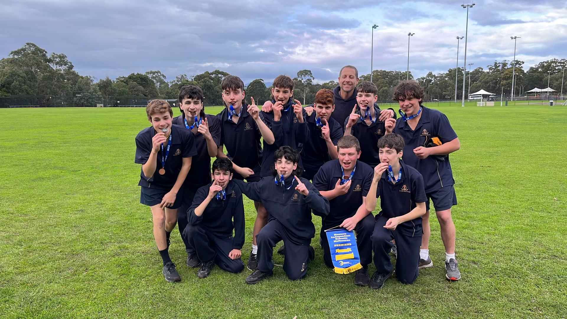 The Year 9 boys' softball team are smiling widely, grinning at the camera as they hold up or bite their gold medals. They are the 2023 EISM Softball champions.
