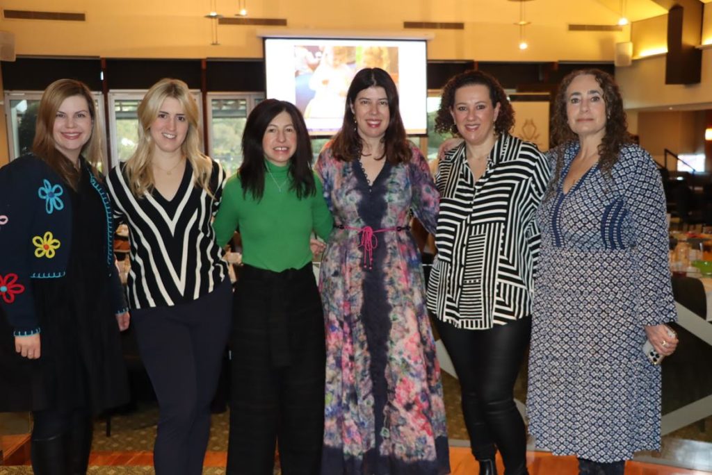 Six women are looking at the camera, smiling. They are dressed in nice clothes. They are at the KDSPA annual breakfast