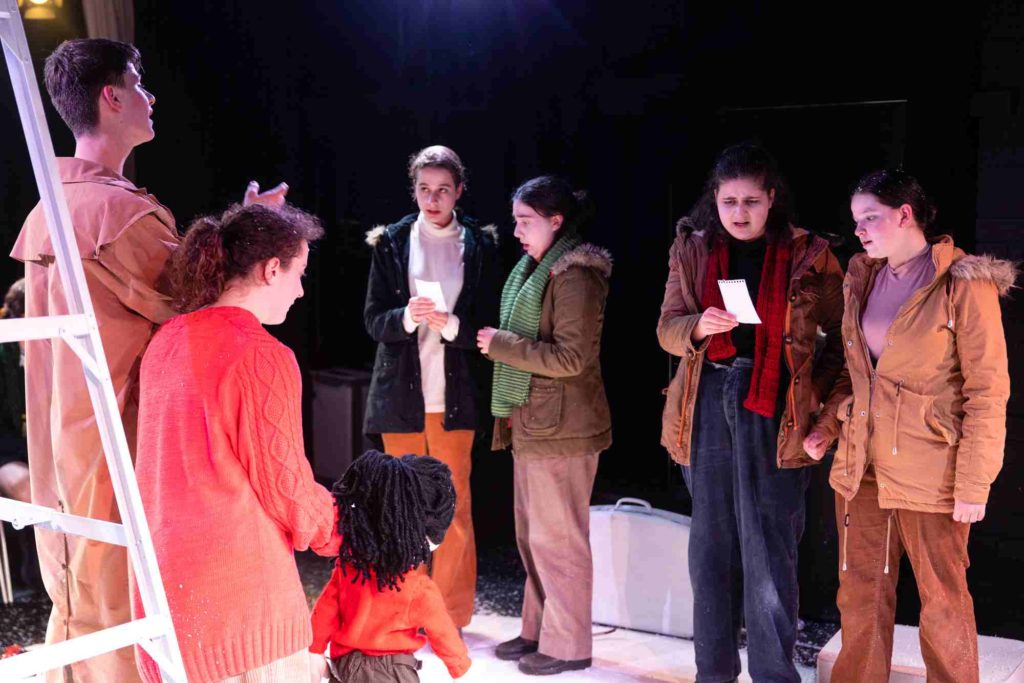 Seven VCE students are on stage in their production of The Snow - they are in costume dressed for snowy weather. There is a ladder on the stage. They are looking down and talking to each other trying to make plans.