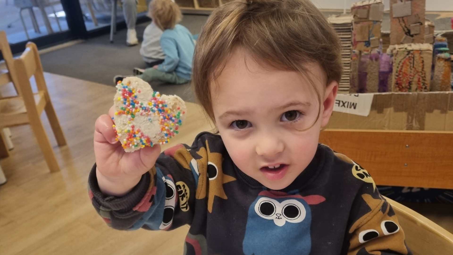 A little learner in our ELC is looking at the camera and holding up heart-shaped bread with sprinkles on it. They are celebrating Tu b'Av