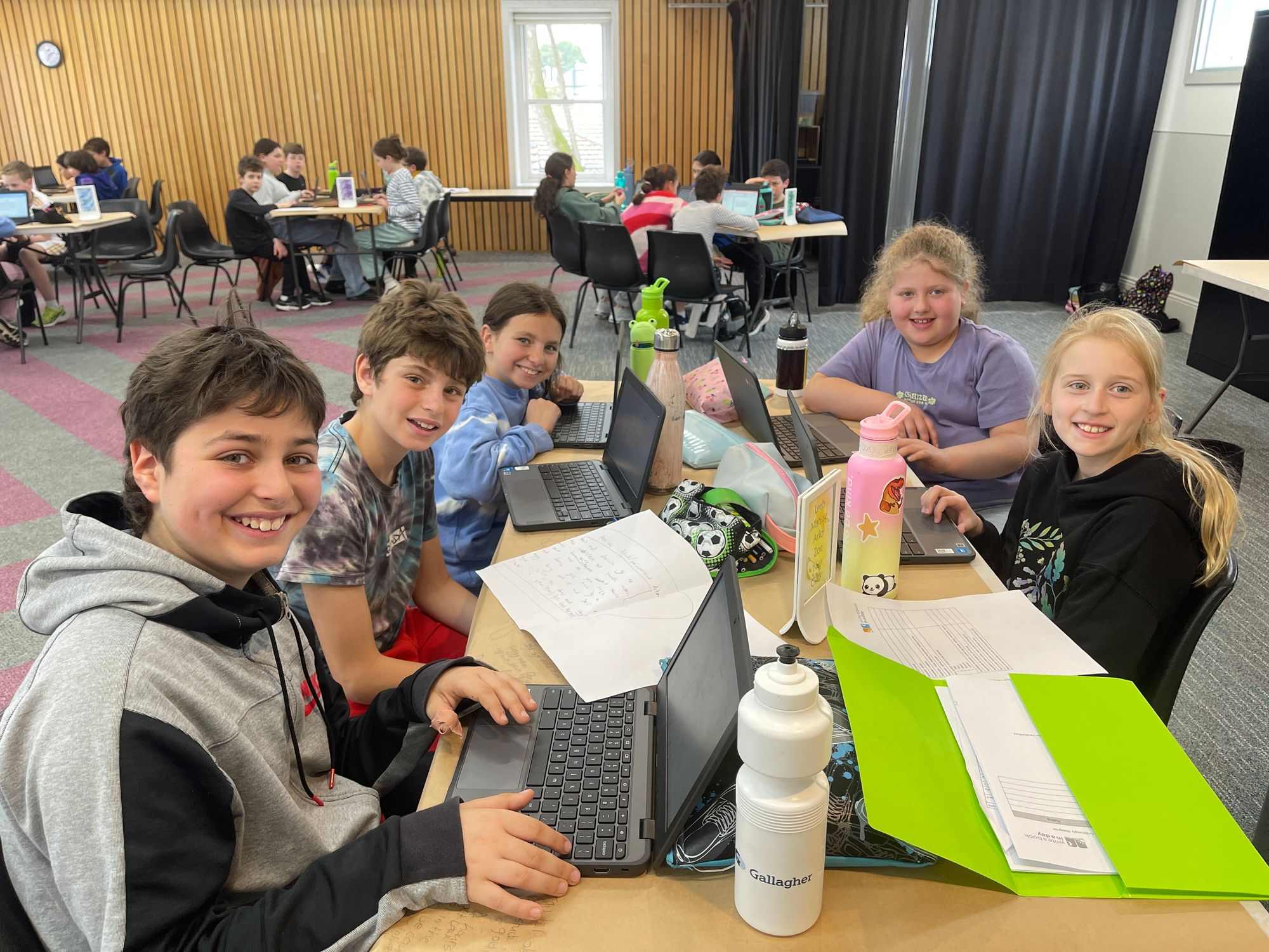 Year 5 students are writing a book in a day! They have laptops and pens and paper in front of them. They are in a small group at desks, smiling at the camera.