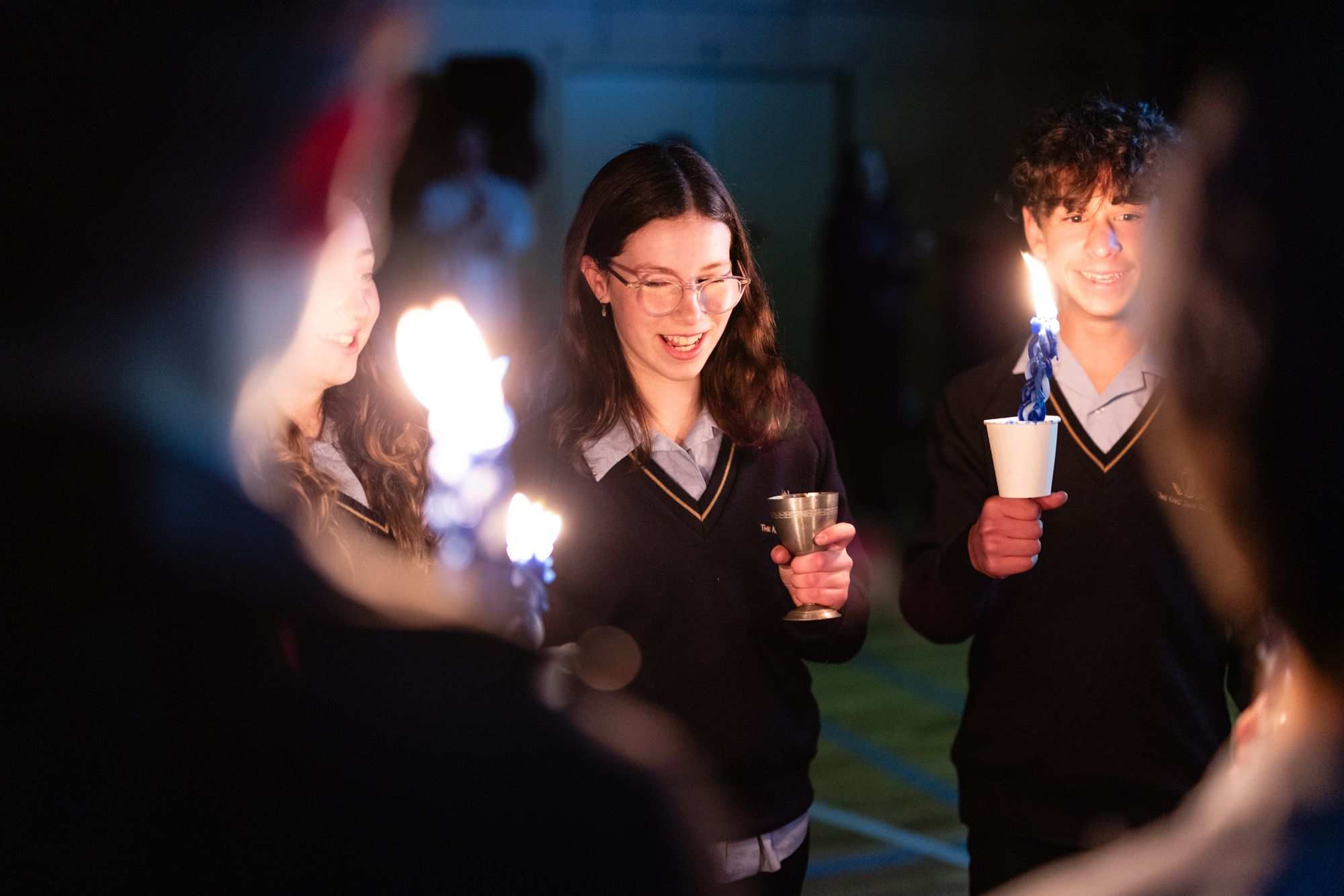 Five Year 7 students are doing Havdalah at the end of Kabbalat Mitzvah. They are holding blue and white braided havdallah candles. Their faces are lit by candlelight