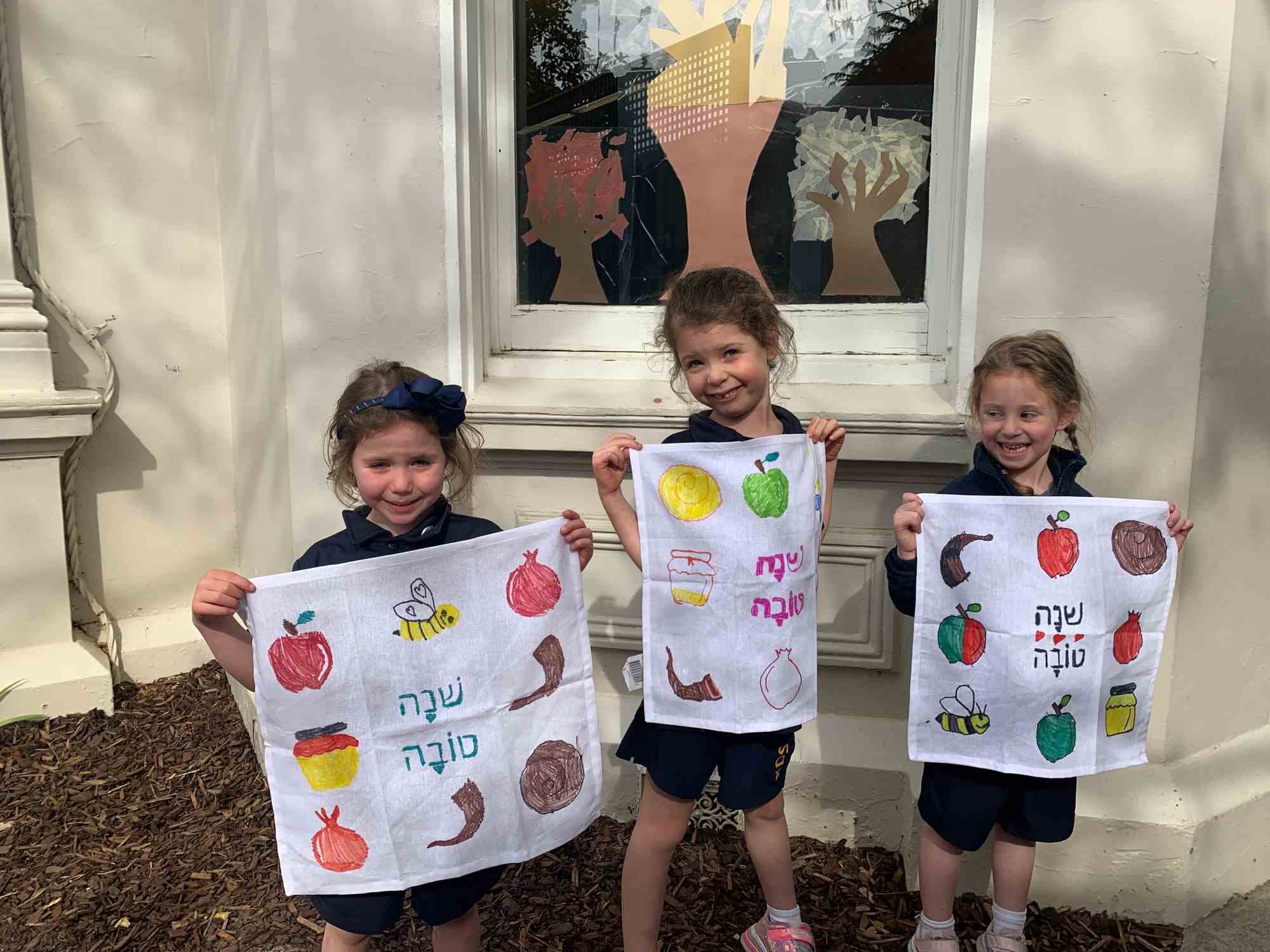 Junior School students are holding tea towels they have painted with Rosh HaShanah symbols and greetings - they say Shana Tova. The three students are smiling and squinting in the sun.