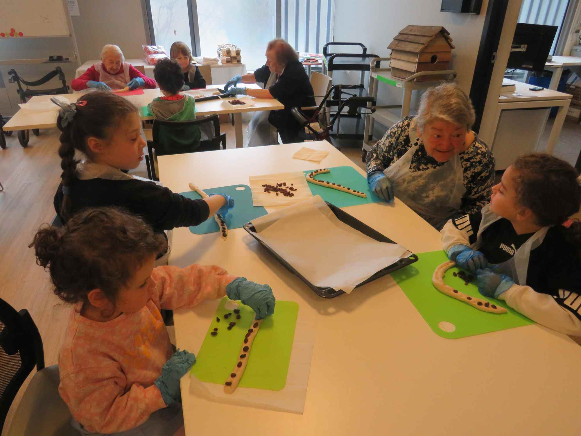 Three KDS ELC students sit with an older person at Emmy Monash. They are crafting at a table. The older person is facing on of the little learners, talking to her.