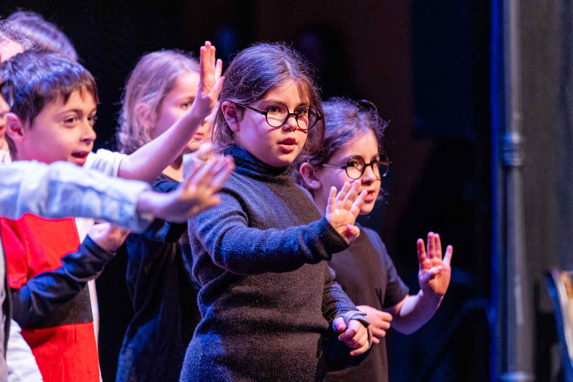 Years 3-5 students are on stage, they are wearing round glasses and dancing with their hands out in front of them.