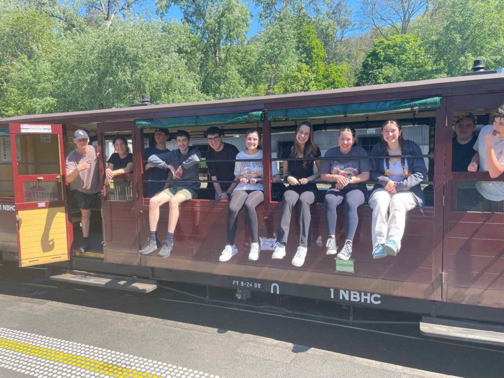 A group of Tourism students are sitting on the Puffing Billy train, they are smiling and waving. They have been investigating sustainable tourism.