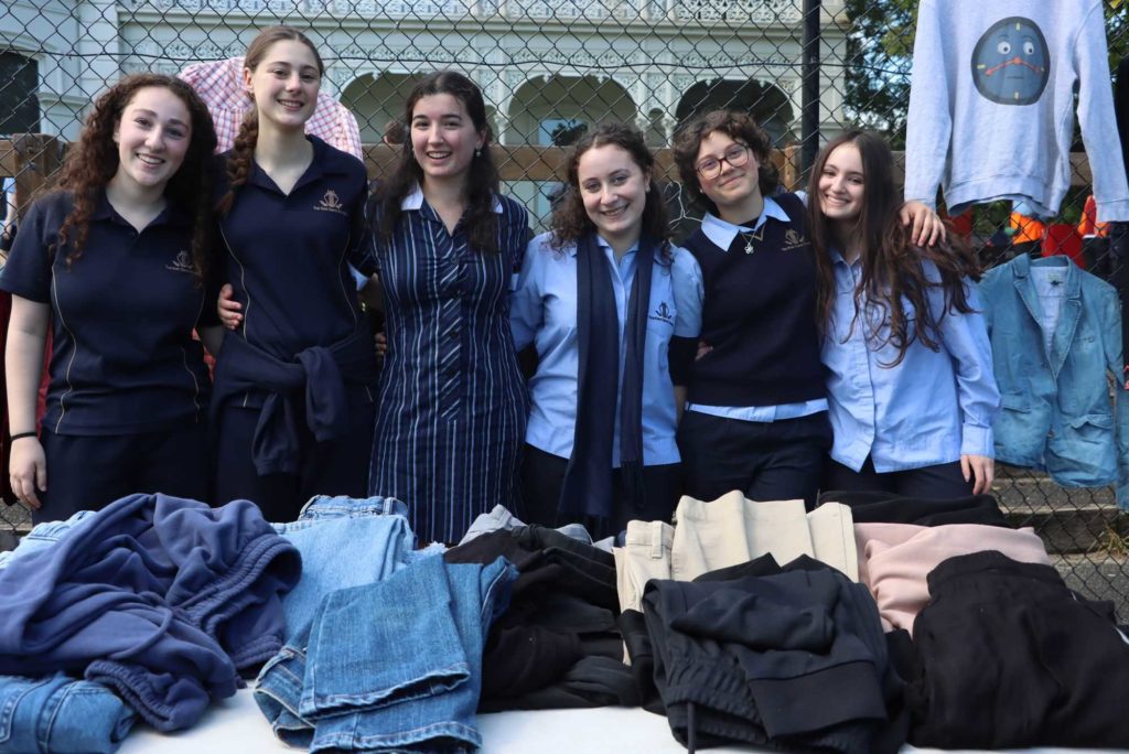 Students look at the camera, smiling. They are behind a table filled with second hand clothes. They are at a ReGen Market to promote sustainability.