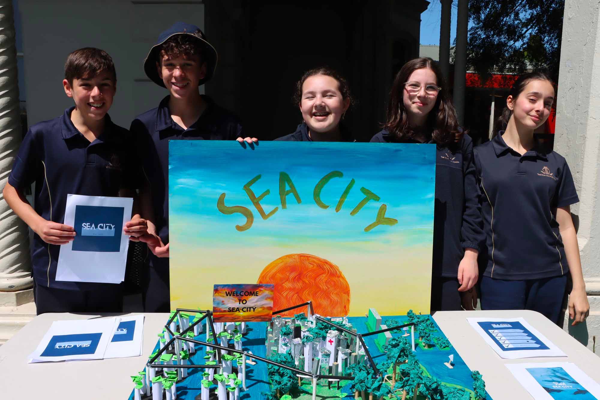 Five students are smiling at the camera. They are standing behind their model of a future sustainable city called Sea City.
