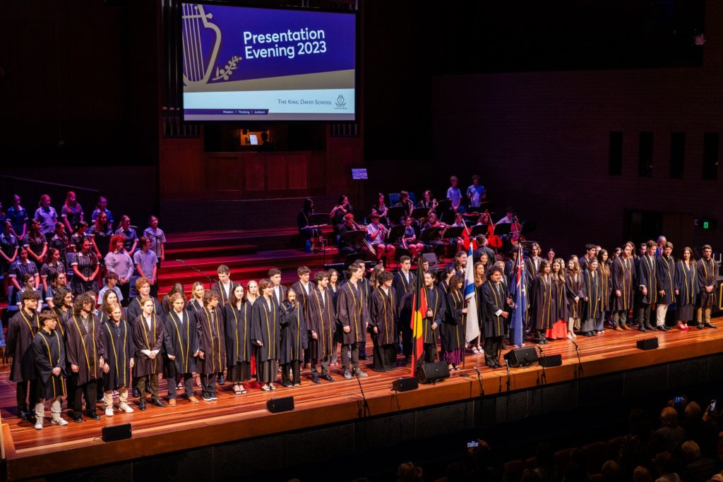 The Class of 2023 is standing on stage, they are celebrating themselves and our Year 6-11 students at Presentation Evening. They are wearing graduation gowns.