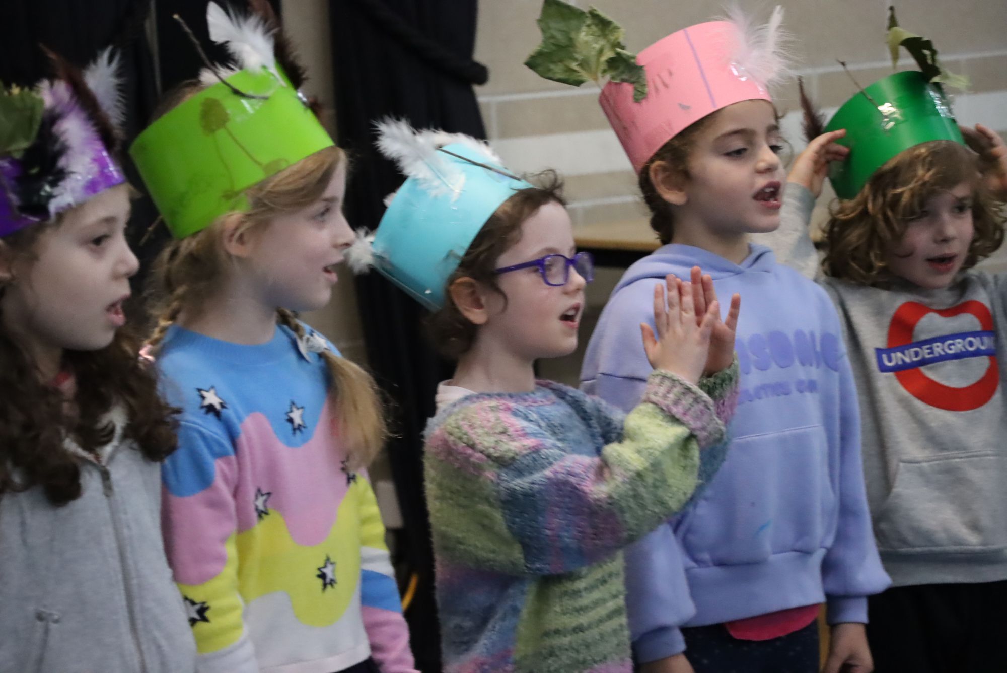 Senior Kinder students are singing at their end of year celebration. They are wearing colourful paper hats.