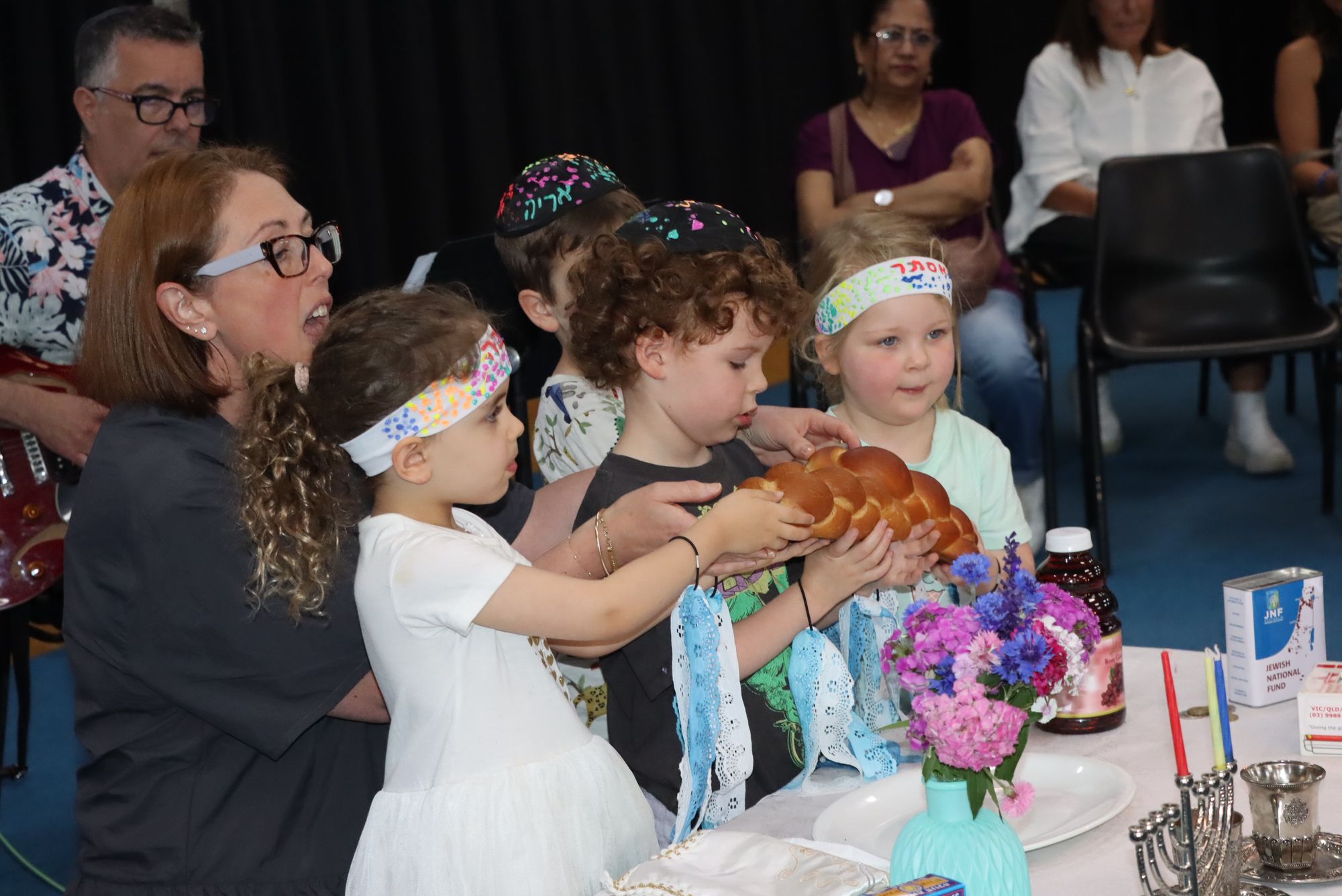 Junior Kinder students are celebrating their end-of-year Shabbat. They are holding challah and smiling near grape juice and Shabbat candles