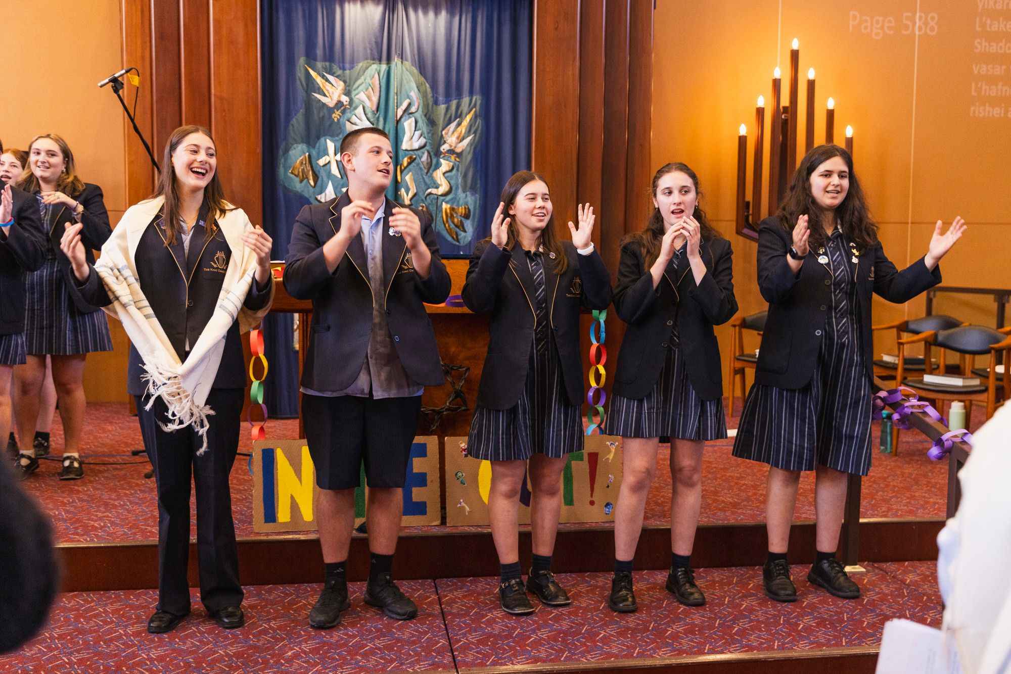 Year 12 students are on the bimah for the 2024 Back to School Service at TBI syngagogue. They are wearing school uniform. One girl is wearing a tallit.