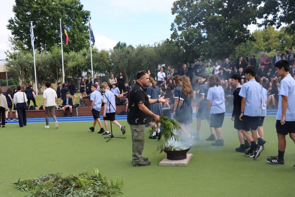 Uncle William Pepper is conducting a Smoking Ceremony to mark the start of the 2024 school year. There is smoke coming from a fire of gum leaves. Students walk past and through the smoke.