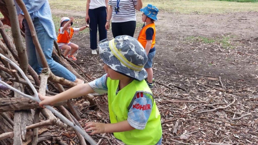 An ELC student in a high-vis vest and a hat plays with a Tee Pee made of sticks. He is at the Urban Forst Bush Kinder with his Gan Rimon KDS class.