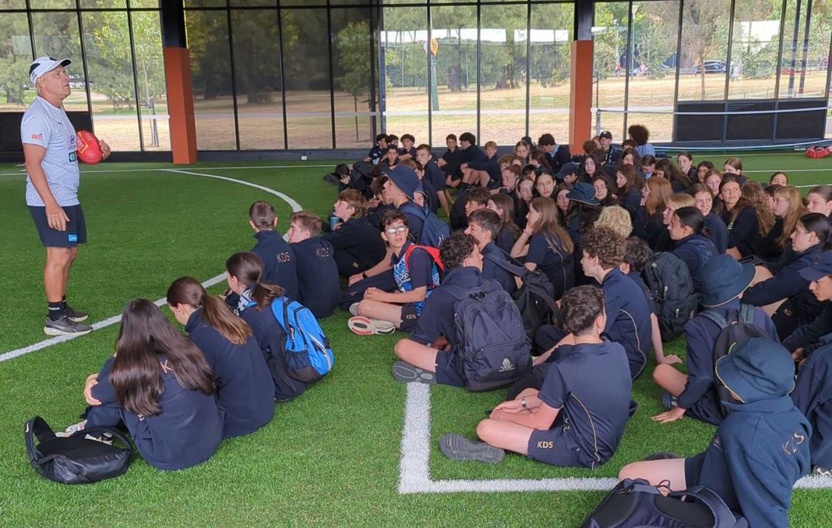 Greg Diesel Williams of Carlton Football Club addresses Year 8 and Year 9 King David students on a oval. He is holding a red ball.