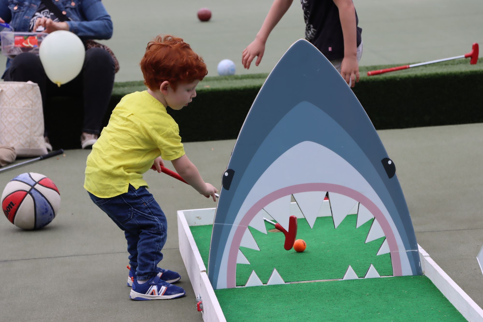 A boy with red hair and a yellow t-shirt plays mini-golf on a shark-themed putting green. He is pictured side on.