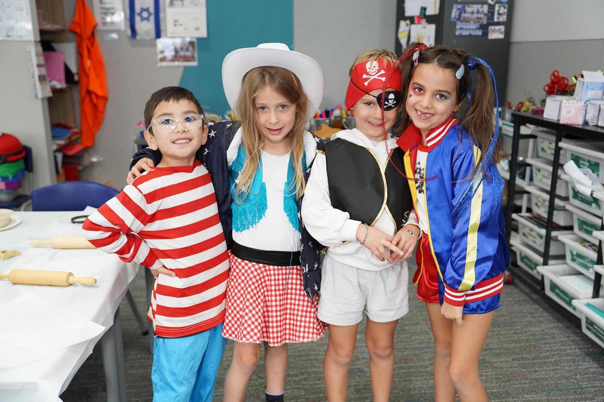 Children dressed in costume as Where's Wally, a Cowgirl, a Pirate and a Cheerleader in shorts smile at the camera. They are celebrating Purim.