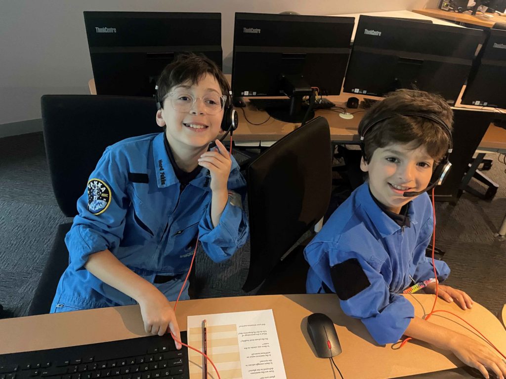 Two Year 5 students are wearing blue Space overalls, as though they work at an International Space Centre. They are wearing headsets and sitting in front of computers at the Victorian Space Science Education Centre (VSSEC)