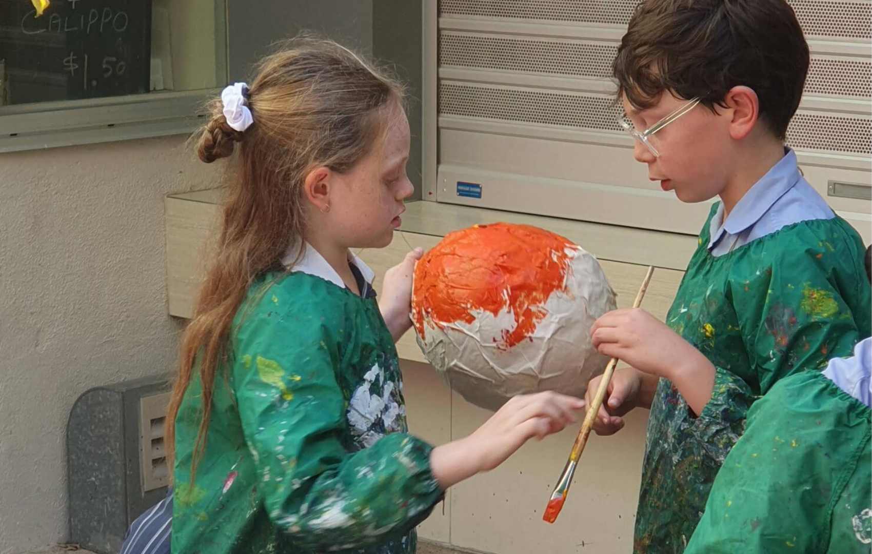 Two Year 5 students are painting a paper mache planet rust red. They are wearing green art smocks.